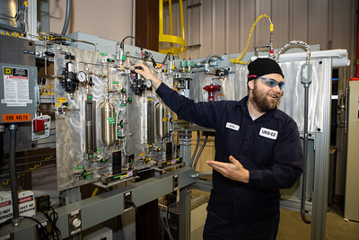 A man in a dark blue jumpsuit standing near and motioning to a large machine.