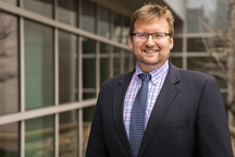 Brian Anderson, a Caucasian man with light brown hair, standing to the right of large, square glass windows while wearing a black suit with a light blue shirt and a navy blue tie, and wearing black-rimmed glasses.