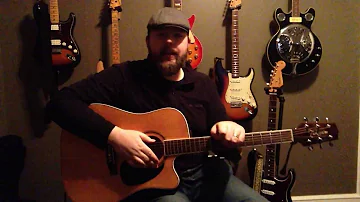 A photograph of a Caucasian man with a dark brown beard, a long sleeve black shirt, and a  grey Kangol hat, sitting with an acoustic guitar in his lap. Also pictured are several other guitars in the background, hanging on an olive green wall.