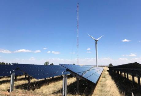 A photograph of various panels of a solar farm, with a bright blue, semi-cloudless sky in the background.