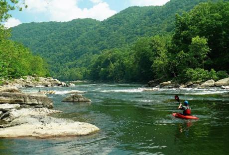 A photograph of a person in a red kayak going down a river with tree-covered hills in the background on a sunny day.